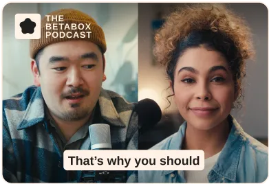 Two podcast hosts, a man wearing a beanie and a woman with curly hair, speaking into microphones.